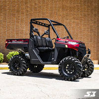 A Polaris Ranger equipped with an S3 Power Sports 3 Bracket Lift Kit, red and black with large rugged tires, is parked on pavement before a tan brick wall. It features two seats, no doors, a roll cage, cargo bed, and green shrubs to the left.