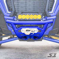 Close-up of an S3 Power Sports 2018+ Polaris Rzr Xp Turbo S Front Winch Bumper, featuring a metallic winch and hook at the center, topped with a bar of six yellow LED lights. Pavement and parts of the grille and suspension are visible.