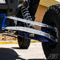 Close-up of S3 Power Sports 2018+ Polaris RZR XP Turbo S HD Billet Aluminum Radius Rods (high clearance lower) installed, with shiny metal arms, coil spring, and off-road tire visible. Sunlight highlights the rugged components against a brick wall.