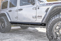A silver Jeep Wrangler Rubicon with large off-road tires is parked on a concrete driveway, featuring S3 Power Sports 2018+ Jeep Wrangler JLU Ambush Series Rock Sliders installed beneath the doors. Jeep and Rubicon logos are visible.