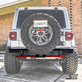 A white Jeep Wrangler JL is parked on a driveway, featuring large off-road tires, a rear-mounted spare tire, and an S3 Power Sports Ambush Series Rear Bumper. A brick building with yellow trim is visible in the background.