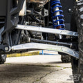 Close-up of a vehicle’s front suspension with S3 Power Sports 2020+ Polaris RZR Pro XP HD Billet Aluminum High Clearance Lower Radius Rods, blue coil spring, metallic control arms, rugged off-road tires, and visible undercarriage on concrete.