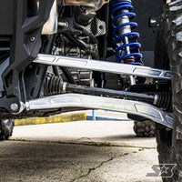 Close-up of a vehicle’s front suspension with S3 Power Sports 2020+ Polaris RZR Pro XP HD Billet Aluminum High Clearance Lower Radius Rods, blue coil spring, metallic control arms, rugged off-road tires, and visible undercarriage on concrete.