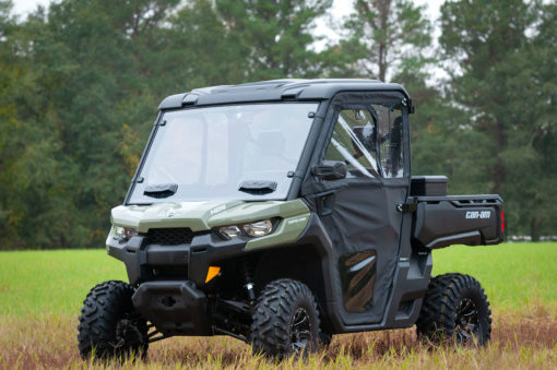 A green and black UTV with a Can-Am Defender Framed Door Kit (2016+) features clear windows, a partially open door, rugged tires, and a short truck bed. It sits on grassy ground before tall trees.