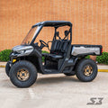 A Can-Am Defender XT with an S3 Power Sports 2 Forward High Clearance A-Arm Kit, featuring large off-road tires, is parked on concrete in front of an orange brick wall with greenery. The open-cabin UTV has visible seats and a rear cargo bed.