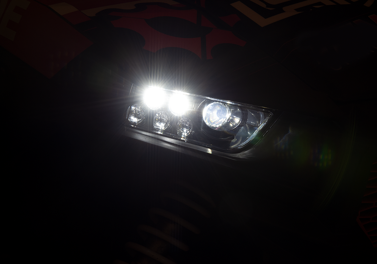 Close-up of a car’s headlight illuminated in the dark. The headlight shows multiple bright beams and reflective elements, casting a strong light that creates lens flares. The rest of the car is mostly hidden in shadow, with faint patterns visible.
