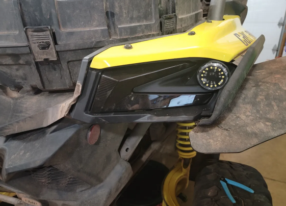Close-up of a yellow ATV, showcasing the Can Am X3 Reverse Light Plug and Play Kit installed as a round black light in the right front headlight. The vehicle is slightly dirty, with visible suspension and part of the front wheel below.