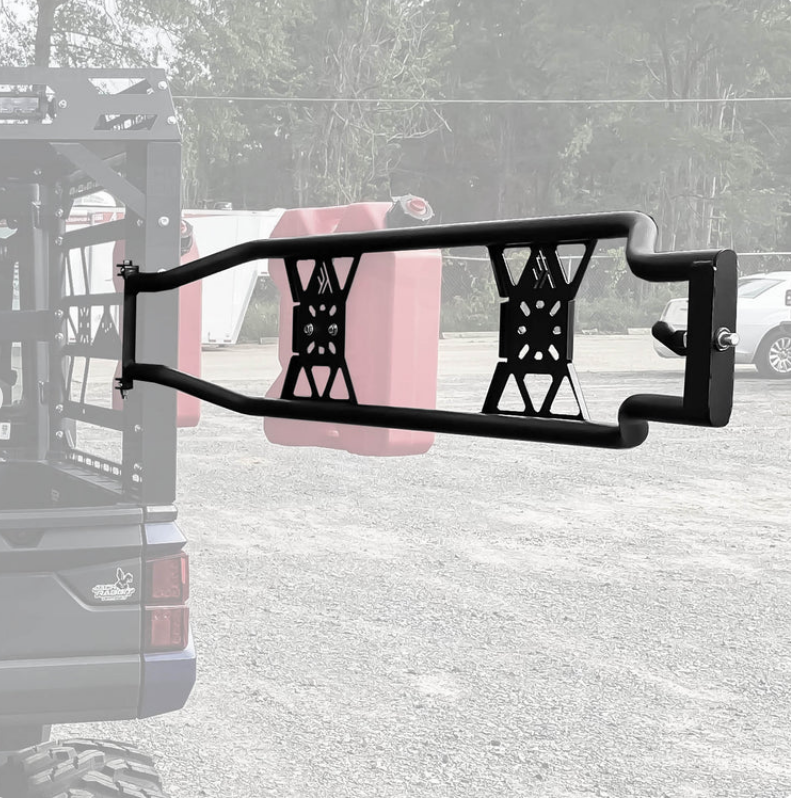 A black REAR TAILGATE for the Thumper Fab Ranger Ultimate Bed Rack is mounted on an off-road vehicle, featuring an X-shaped support and holding two red fuel cans. In the background are trees, a gravel lot, and a partially visible vehicle.
