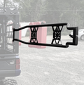 A black REAR TAILGATE for the Thumper Fab Ranger Ultimate Bed Rack is mounted on an off-road vehicle, featuring an X-shaped support and holding two red fuel cans. In the background are trees, a gravel lot, and a partially visible vehicle.