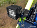 Close-up of a square LED light attached with 1.75 Cage Light Mounting Brackets on a neon yellow off-road vehicle frame; black bracket, partial windshield, blue seat, and green foliage in background are visible.