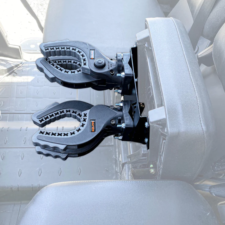 The 2017+ Can-Am Defender Hidden Rifle Mount features two black ratcheting holders with rubber X-cutout grips and adjustment knobs, mounted side-by-side on the back of a vehicle seat to securely hold rifles or equipment.