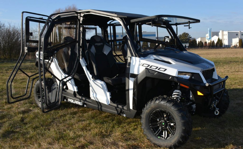 A Polaris General 1000-4 Hard Cab Enclosure with open doors is parked on grassy terrain. This off-road utility vehicle features large rugged tires, a roll cage, seating for at least four, and industrial buildings plus trees in the background.