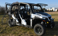 A Polaris General 1000-4 Hard Cab Enclosure with open doors is parked on grassy terrain. This off-road utility vehicle features large rugged tires, a roll cage, seating for at least four, and industrial buildings plus trees in the background.