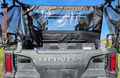 Close-up of a dusty Honda Pioneer 1000-5 with the Soft Center Panel, showing the embossed tailgate, two red taillights, mesh side panels, and clear rear window. Blue sky and fields appear in the background.