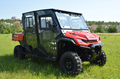 A Honda Pioneer 1000-6 Full HardCab Enclosure (2023–Current) in red, with four doors, large rugged tires, black detailing, and transparent windows, is parked on green grass under a clear sky with houses and trees in the background.