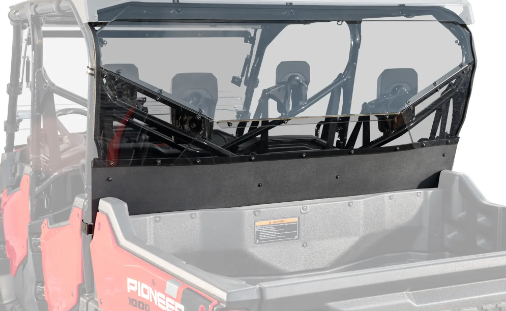 A close-up of the Honda Pioneer 1000-6 Rear Windshield shows its clear, tinted, angular design with a black lower panel mounted on a red UTV’s cargo bed, with seats and headrests visible through the glass.