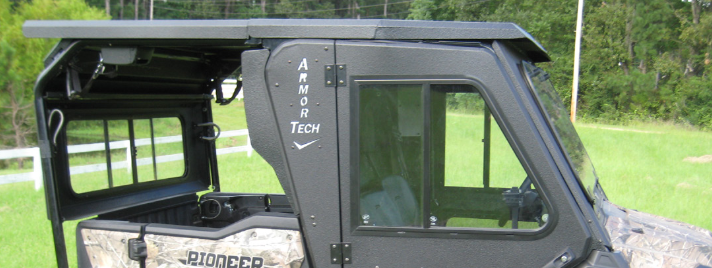 Side view of the Pioneer 1000-6 Roof Armor Tech utility vehicle with a matte black cab enclosure and tinted windows, parked on green grass near a white fence and trees. PIONEER and ARMOR TECH logos are visible on its exterior.