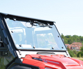 A close-up of a Honda Pioneer 1000 Windshield Kit installed on a red vehicle, with sky reflections and Dual Clutch Transmission on the hood. Blurred trees and houses appear in the background beneath a clear sky.