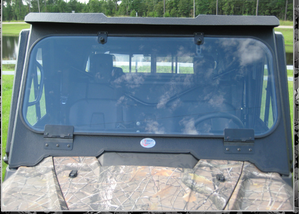 A Honda Pioneer 1000/1000-5/1000-6 with a Glass Flip Out Windshield Armor Tech (2022+) in camouflage sits on grassy ground near a pond and trees; its large tinted windshield reflects sky and clouds with the interior faintly visible through the glass.
