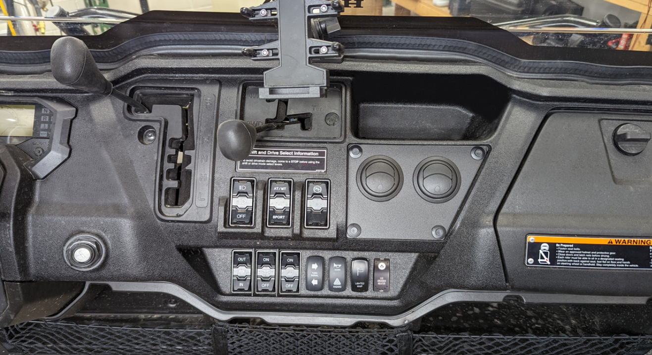 The image shows the dashboard of a Honda Pioneer 1000 (2022-2024) with an Ice Crusher Heater installed, multiple toggle switches, round dials, a gear shift lever, a central device holder, warning label on the right, and storage net below.