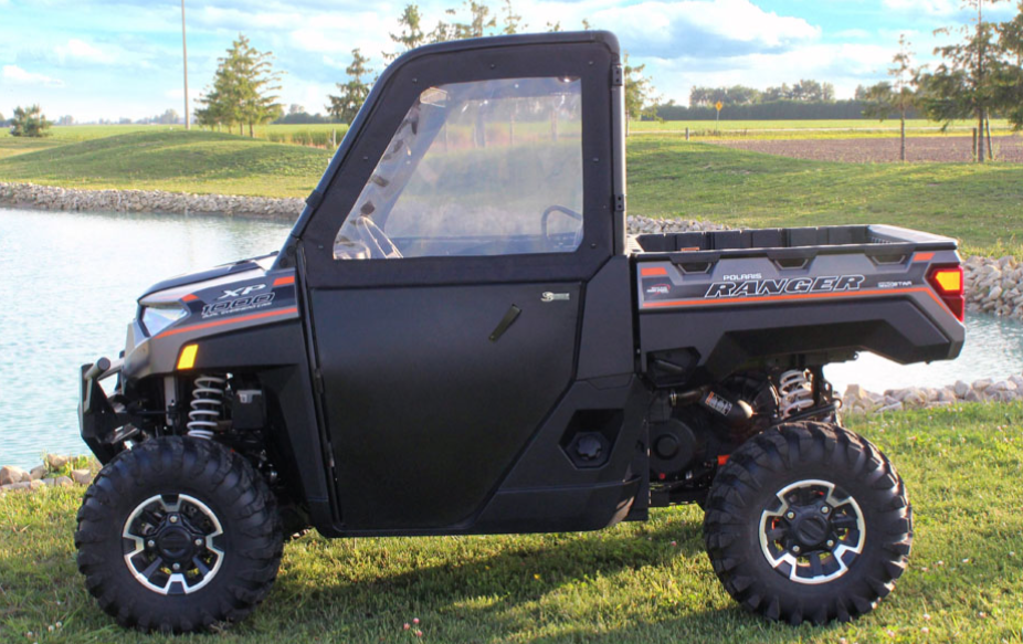 A Polaris Ranger 1000 with Convertible Doors, featuring a side enclosure and large off-road tires, is parked on grass near a pond with trees and a clear sky in the background, highlighting its rugged styling.