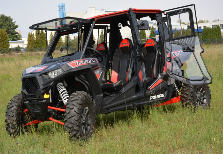 A Polaris RZR 1000-4 Turbo with a Hard Cab Enclosure, finished in black and red, is parked on grassy ground; trees and industrial buildings are blurred in the background.