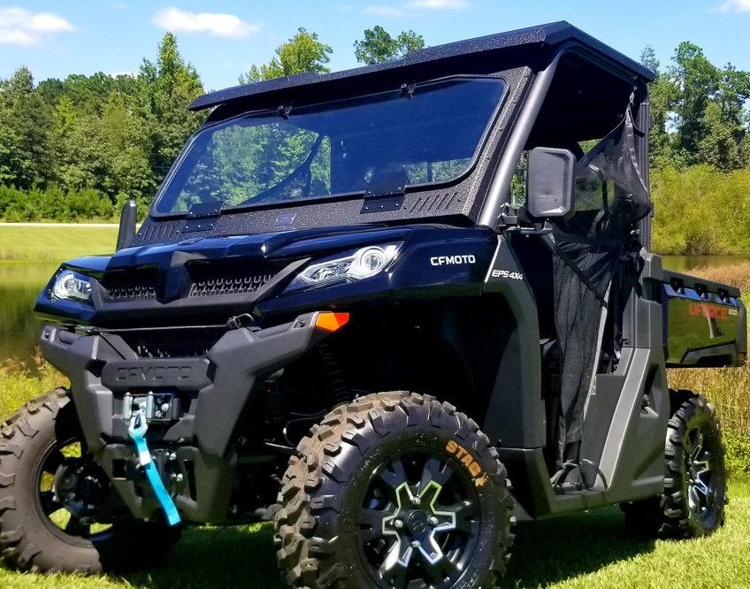 A CFMoto UForce 1000/1000XL with a Glass Flip Down Windshield Armor Tech is parked on grass near a pond, featuring rugged tires, side doors, and a front winch. Trees and blue sky in the background suggest a sunny outdoor setting.
