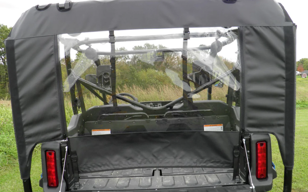 Rear view of a Honda Pioneer 1000-5 with a Soft Back Panel featuring a clear plastic rear window, revealing the interior seats, steering wheel, and dashboard. The vehicle is parked on grass with trees and tall grass behind it.