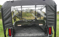 Rear view of a Honda Pioneer 1000-5 with a Soft Back Panel featuring a clear plastic rear window, revealing the interior seats, steering wheel, and dashboard. The vehicle is parked on grass with trees and tall grass behind it.