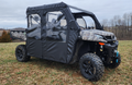 A CF Moto UForce 1000 XL Crew with Soft Doors in camouflage sits on grass, featuring a black weatherproof cabin enclosure, clear vinyl windows, and large rugged tires. Leafless trees and cloudy skies are visible in the background.