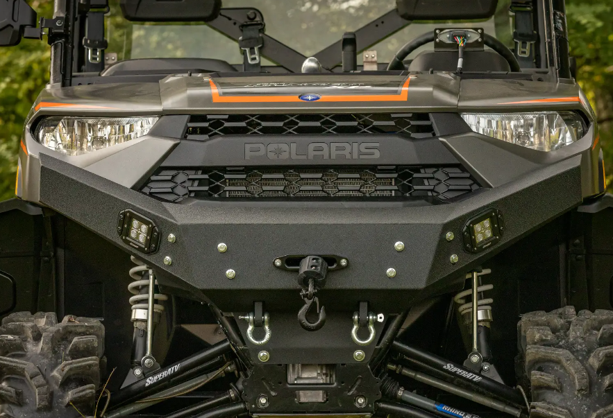 Close-up of a Polaris Ranger XP 1000 Winch Ready Front Bumper featuring the grille with Polaris logo, headlights, rugged bumper with mounted winch, suspension parts, large tires, and LED fog lights against a blurred outdoor background.