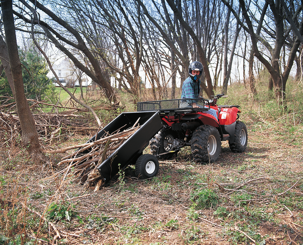 A person wearing a helmet drives a red ATV with a black dump trailer attached, unloading branches in a wooded area surrounded by leafless trees and scattered debris. The ground is covered with dry leaves, twigs, and green undergrowth.