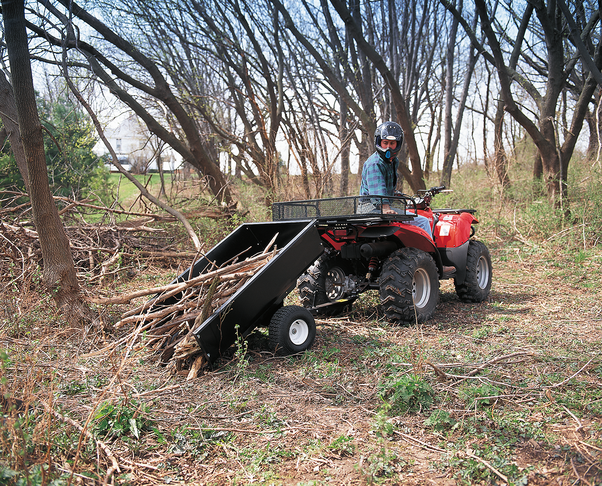 A person wearing a helmet drives a red ATV with a black dump trailer attached, unloading branches in a wooded area surrounded by leafless trees and scattered debris. The ground is covered with dry leaves, twigs, and green undergrowth.