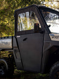 A close-up of Polaris Ranger 570sp Convertible Doors shows a black panel with a clear plastic window on the vehicles side, parked outdoors among green trees with natural sunlight filtering through the foliage.