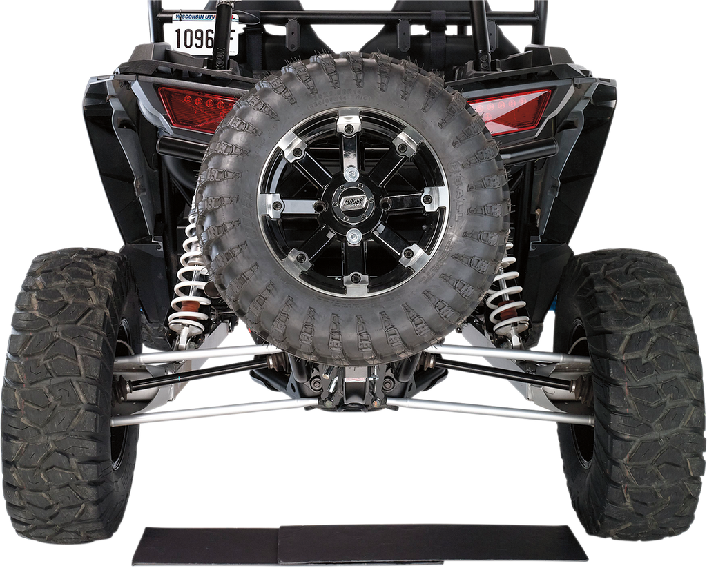 Rear view of an off-road vehicle showing its large, rugged spare tire mounted on the back, flanked by two thick rear tires with deep treads. Visible are the suspension coils, tail lights, license plate, and part of the undercarriage against a white backgr