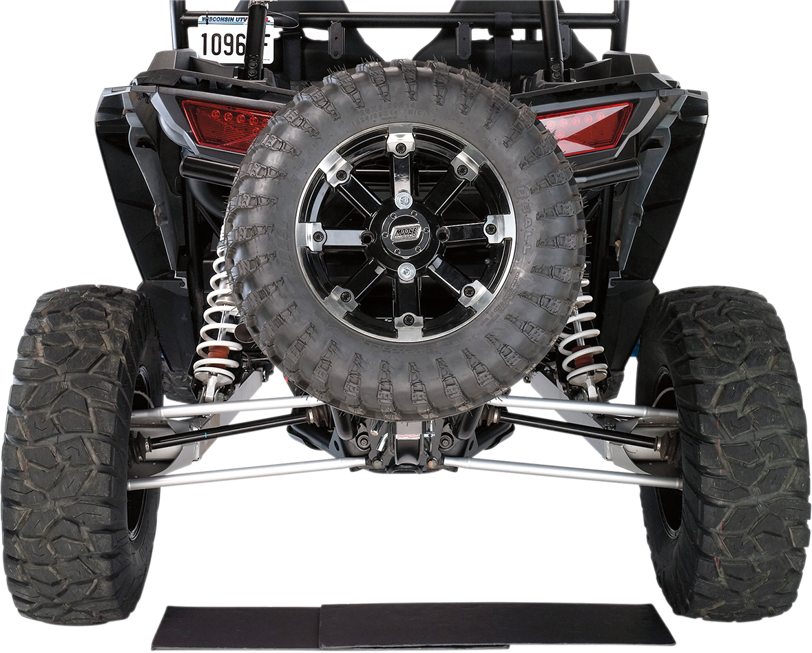 Rear view of an off-road vehicle showing its large, rugged spare tire mounted on the back, flanked by two thick rear tires with deep treads. Visible are the suspension coils, tail lights, license plate, and part of the undercarriage against a white backgr