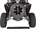 Rear view of an off-road vehicle with large, rugged tires, prominent exposed suspension springs, and a spare tire mounted in the center. Two red taillights, a partial license plate, and sturdy metal axles are visible against a white background.