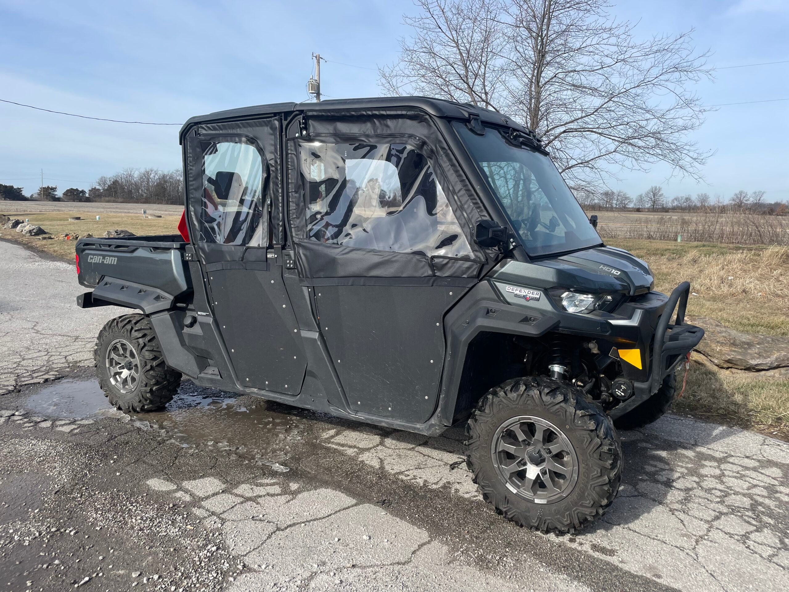 A Can-Am Defender fitted with Max Framed Upper Soft Doors for SuperATV lower doors is parked on cracked asphalt, surrounded by dry grass and leafless trees under a partly cloudy sky in a rural area.