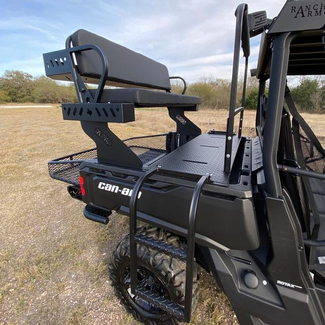 A Can-Am Defender with a Quick Connect High Seat, featuring a rear-mounted bench with backrest elevated above the bed and accessed by metal steps and railings, is parked on grass under partly cloudy skies with trees in the background.