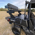 A Can-Am Defender with a Quick Connect High Seat, featuring a rear-mounted bench with backrest elevated above the bed and accessed by metal steps and railings, is parked on grass under partly cloudy skies with trees in the background.