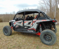 A Polaris Rzr Xp 4 Turbo S - Soft Top with black and red accents is parked on grass among leafless trees, featuring large rugged tires, roll bars, and set against a cloudy sky backdrop.