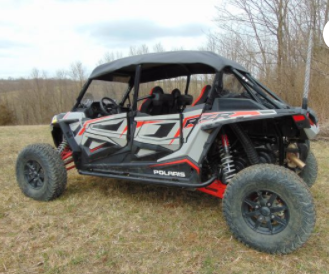 A Polaris Rzr Xp 4 Turbo S - Soft Top with black and red accents is parked on grass among leafless trees, featuring large rugged tires, roll bars, and set against a cloudy sky backdrop.