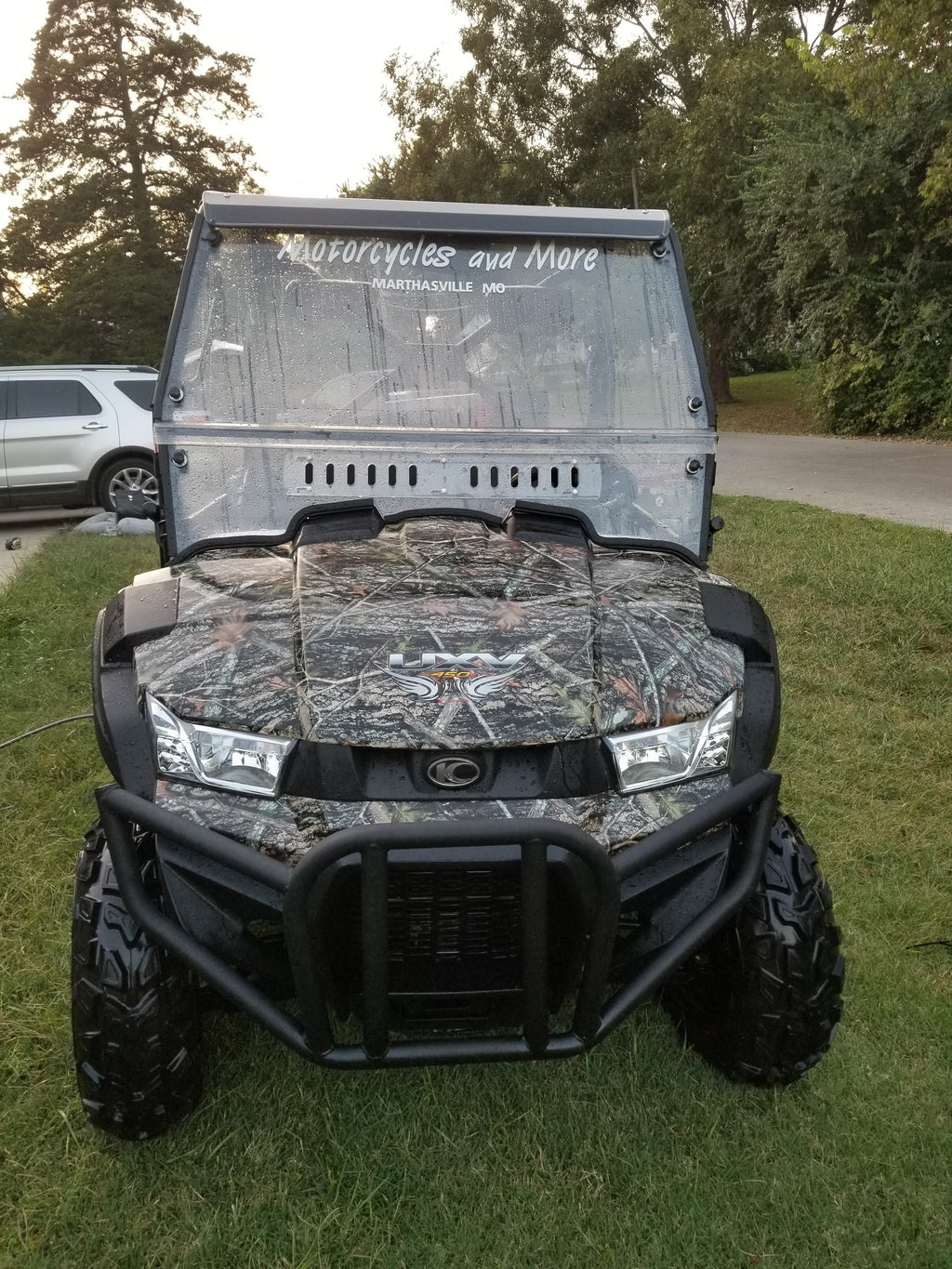 A Kymco Uxv 450I with a Split Lexan Windshield (2015-2018) featuring a camouflage pattern is parked on grass near a road. Motorcycles and More, Marthasville, MO is on the windshield, with a white SUV and trees in the background.