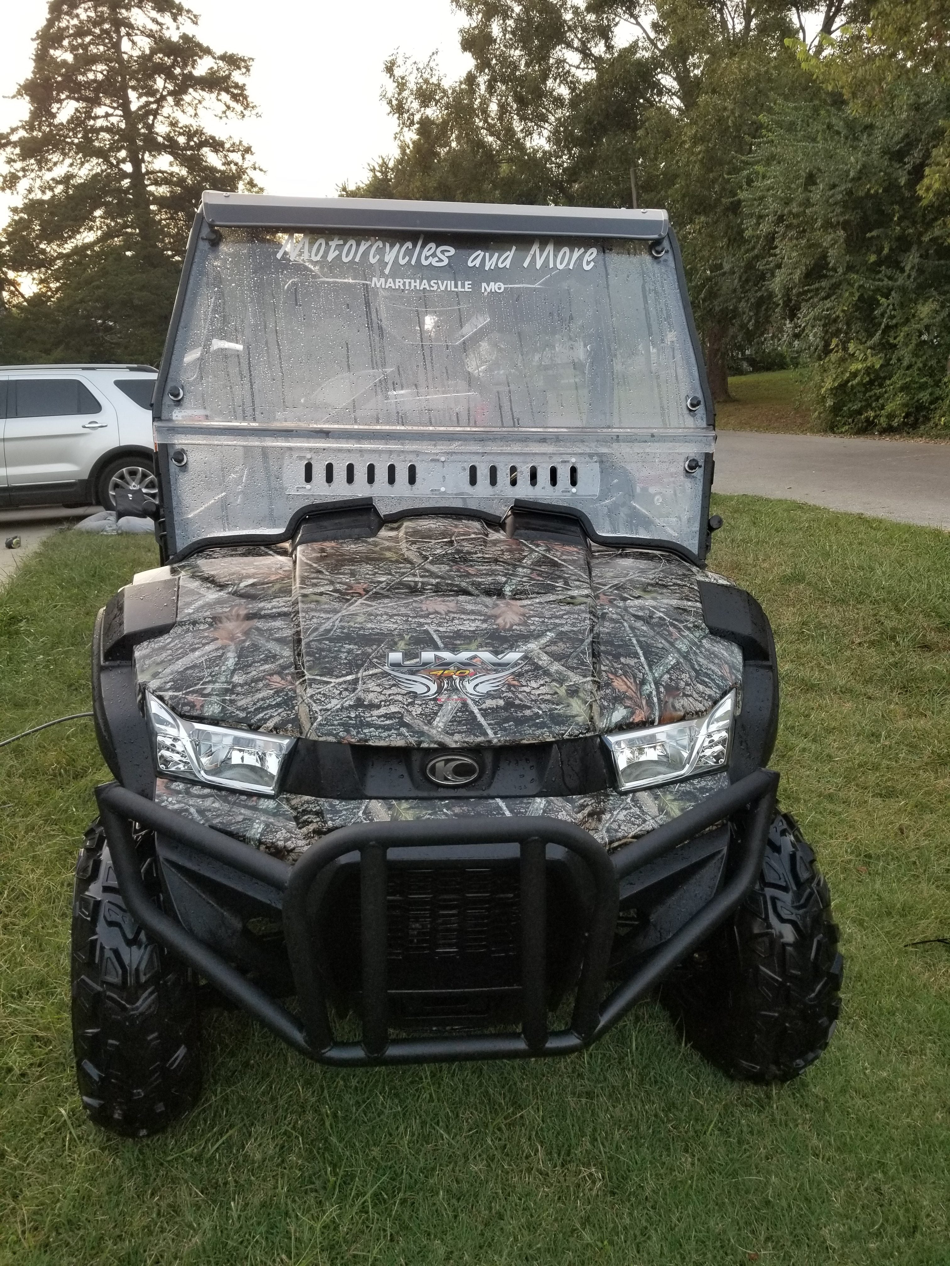 A Kymco Uxv 450I with a Split Lexan Windshield (2015-2018) featuring a camouflage pattern is parked on grass near a road. Motorcycles and More, Marthasville, MO is on the windshield, with a white SUV and trees in the background.