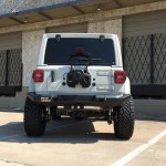 A white Jeep Wrangler with large off-road tires and a 2018+ Jeep JL Rubicon Stealth Rear Bumper (Full Width, Texture Black) is parked by a beige stone building with barred windows and closed gray roll-up doors. The spare tire is removed; rear windows are 