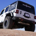 A white Jeep JL Rubicon with large tires is parked on rocky terrain under a blue sky, showcasing its 2018+ Stealth Rear Bumper Mid Width in texture black from behind, along with visible suspension, spare tire, and rear window decals.