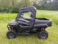 A Honda Pioneer 700 (2014+) with a Soft Door Kit is parked on green grass. The black UTV features large rugged tires, red suspension springs, and clear plastic windows. Trees and tall grass fill the cloudy background.