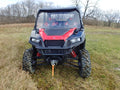 A red and black off-road ATV with large muddy tires features a Polaris General 1 Pc. Lexan Windshield, visible suspension springs, and two seats. It’s parked on grassy terrain under a cloudy sky, with leafless trees in the background.