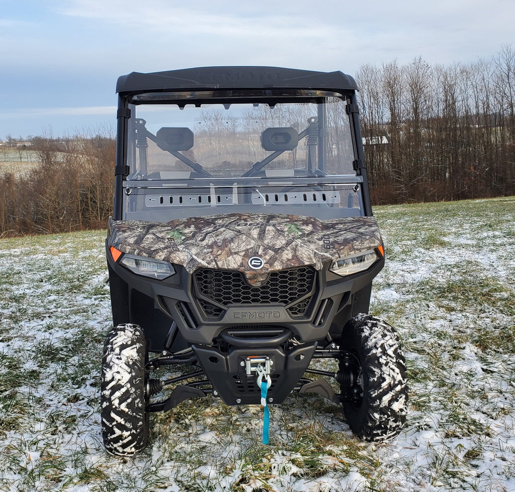 A Cfmoto Uforce 600 featuring a 2 pc windshield with optional hard coat, vents, and clamps is parked on a grassy field with snow patches. It has black and camo colors, large rugged tires, bare trees behind, and a blue tow strap on the front winch.