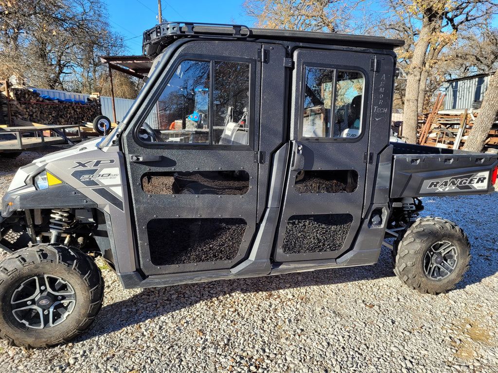A Polaris Ranger Crew XP 1000 with Armor Tech Full Steel Doors is parked on gravel; the vehicle features rugged off-road tires. Leafless trees, stacked logs, and a shed appear in the background on a sunny day.